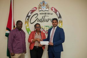 Left, Director of Mashramani, Andrew Tyndall, AMDI Corporate Communications Officer, Alleya Hamilton, handing over the donation to Minister of Culture, Youth and Sports, Hon. Charles Ramson.